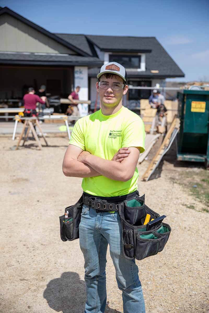 Construction stduent in neon shirt with tool belt stands in front of a house under construction with materials and workers nearby