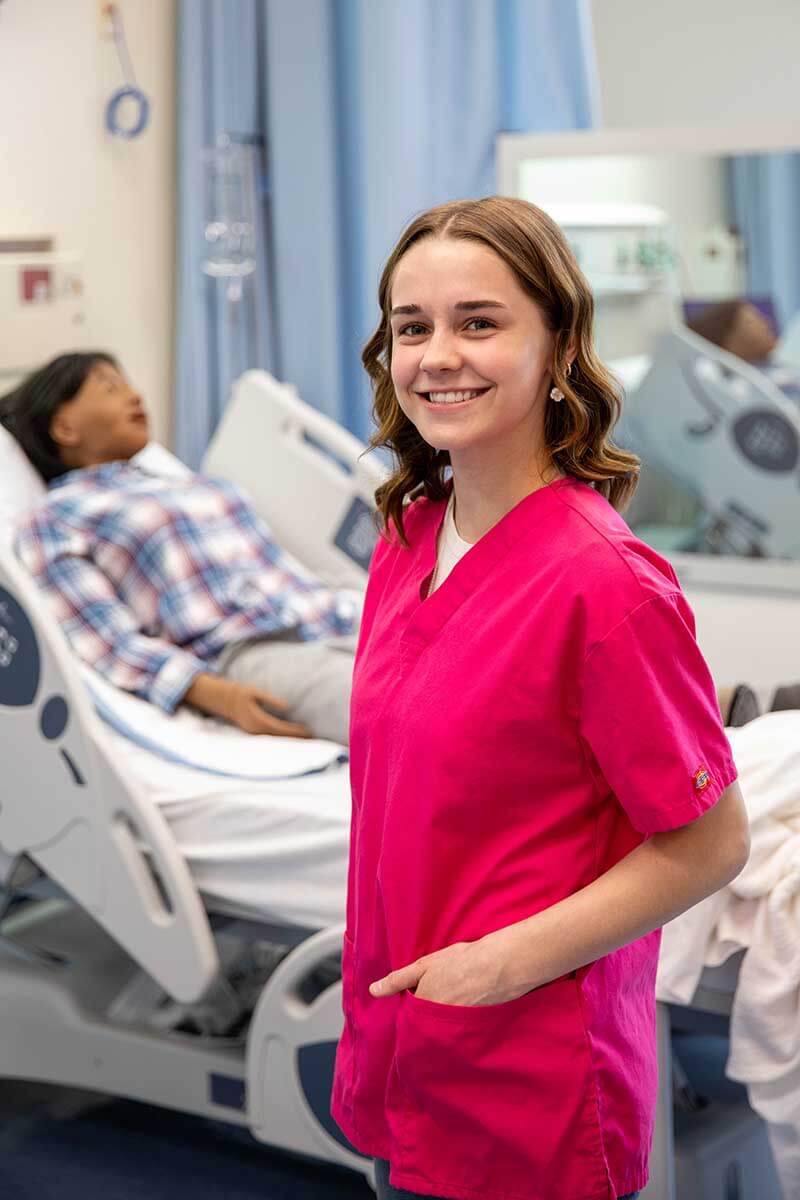 Nursing student in pink scrubs stands beside a patient in a hospital bed with medical equipment and curtains in the background