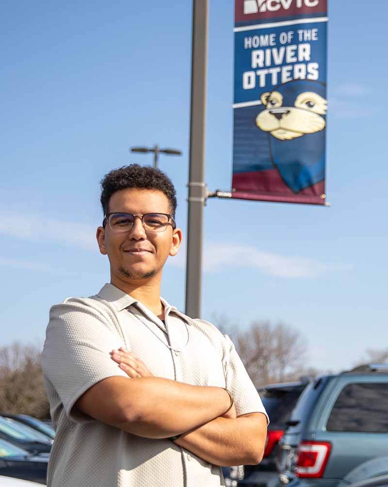Person stands with arms crossed in front of a 'Home of the River Otters' banner with cars and trees in the background