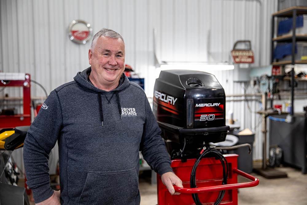 Steve standing in a workshop beside a Mercury 50 outboard motor mounted on a red stand, with tools and equipment visible in the background.
