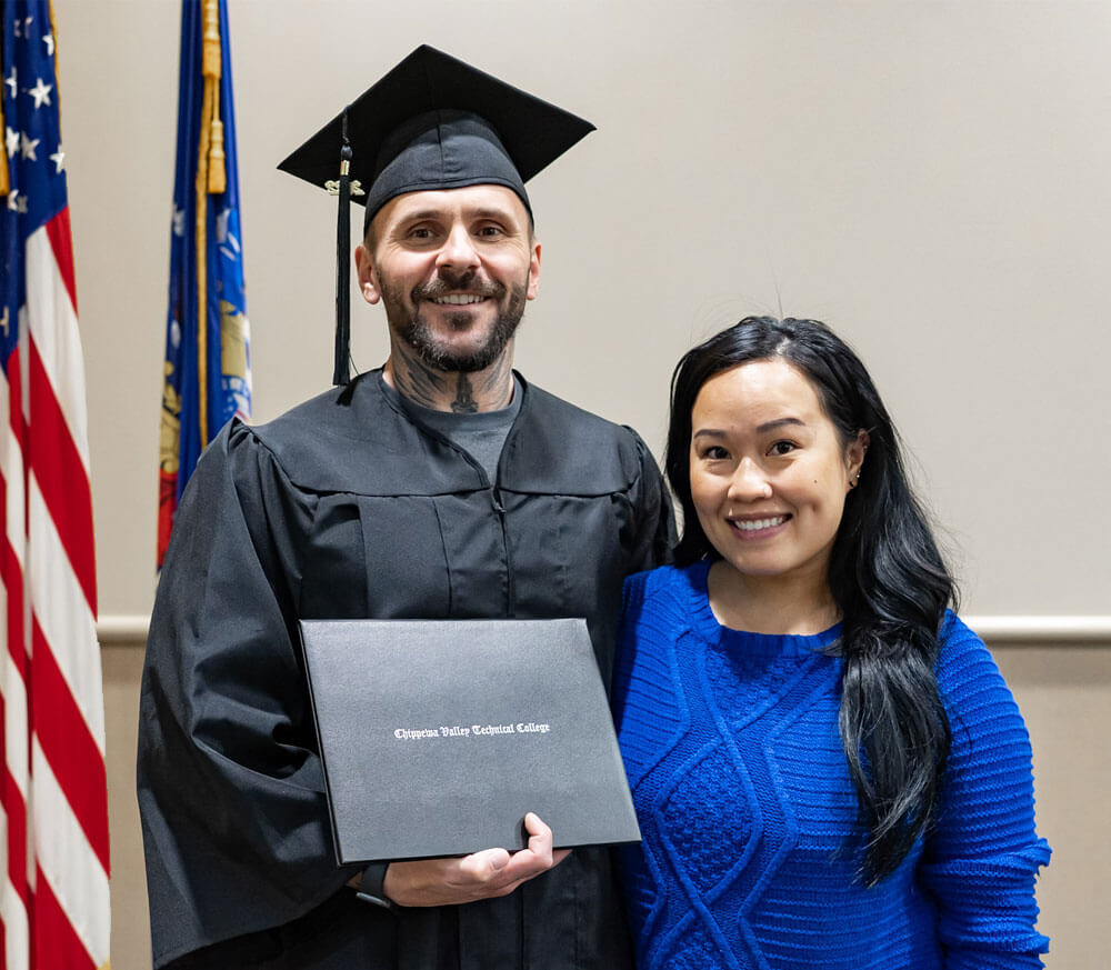 A man on the left in a graduation gown hugging an woman on the right. Both of them are smiling.