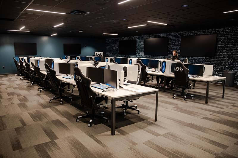 A wide-angled view of a computer classroom with one student standing by one of the workstations