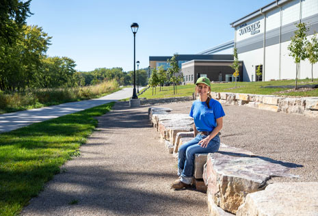 Female in blue shirt and jeans outside sitting on a rock ledge near a walking trail and new event center.